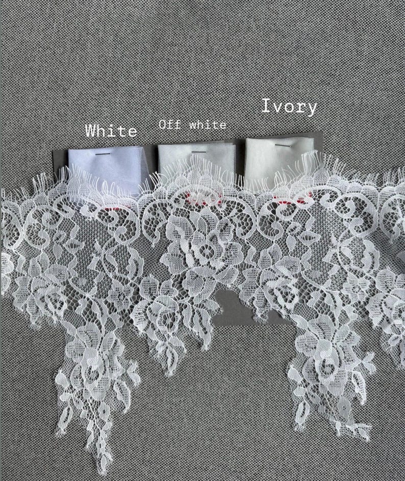 May include: A wide strip of white floral lace trim with scalloped edges, displayed alongside three small fabric swatches labelled White, Off white, and Ivory. The lace features an intricate botanical pattern with delicate mesh detailing.
