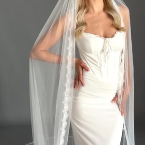 May include: A white bridal gown with a fitted bodice and a long, flowing skirt. The dress has a sweetheart neckline and delicate lace trim. A sheer veil with lace edging drapes over the shoulders.