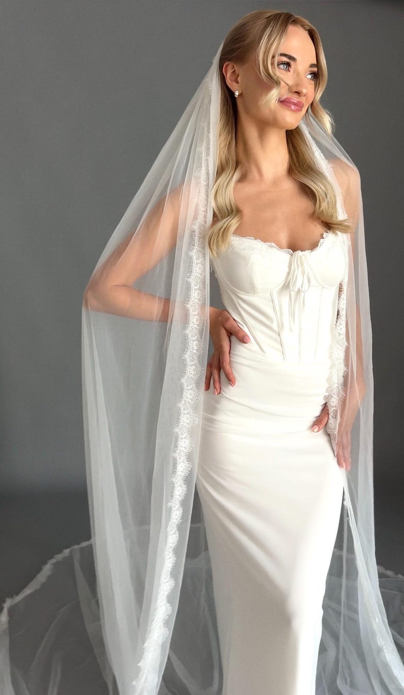May include: A white bridal gown with a fitted bodice and a long, flowing skirt. The dress has a sweetheart neckline and delicate lace trim. A sheer veil with lace edging drapes over the shoulders.