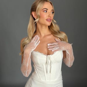 May include: A white bridal gown with a corset-style bodice and a flowing skirt. The dress is paired with delicate lace gloves. The model has blonde hair and is wearing pearl earrings. The background is a solid gray.