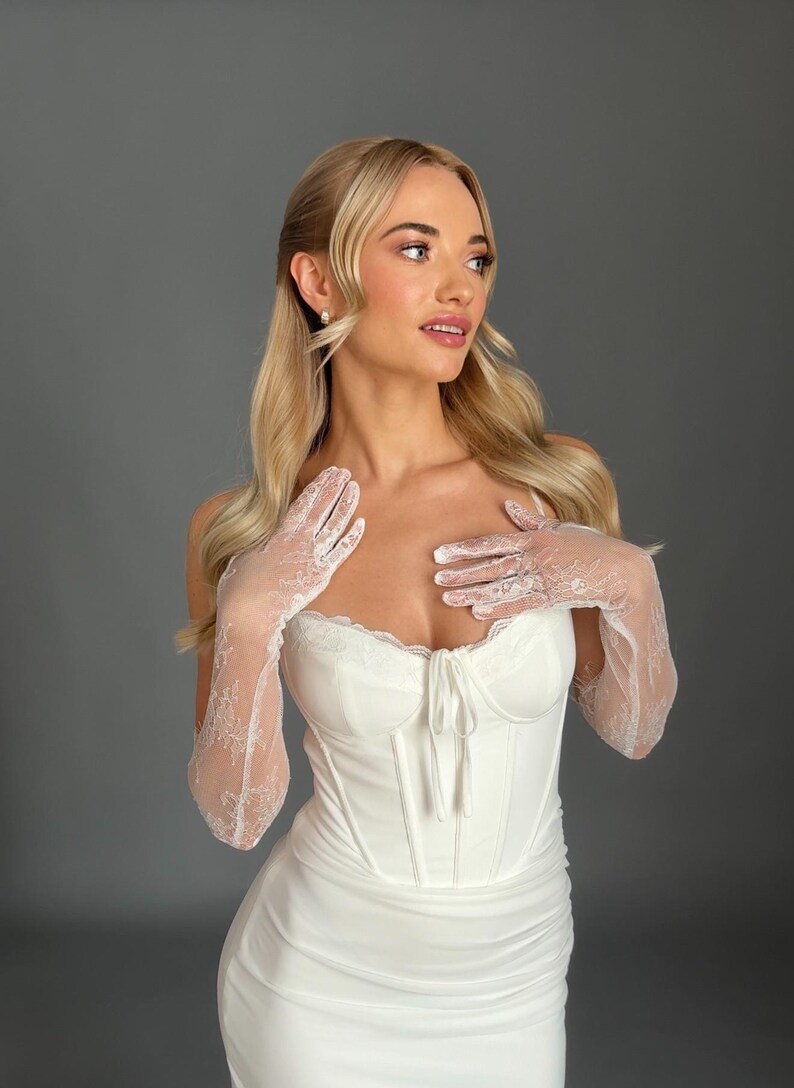 May include: A white bridal gown with a corset-style bodice and a flowing skirt. The dress is paired with delicate lace gloves. The model has blonde hair and is wearing pearl earrings. The background is a solid gray.