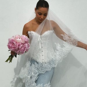 May include: A white strapless corset top with a front button closure, paired with light blue denim jeans. A long, sheer white veil with intricate lace trim edges is draped over the head and shoulders, accompanied by a bouquet of pink peonies.