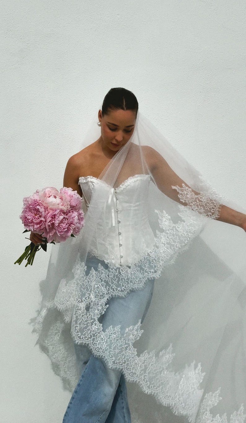 May include: A white strapless corset top with a front button closure, paired with light blue denim jeans. A long, sheer white veil with intricate lace trim edges is draped over the head and shoulders, accompanied by a bouquet of pink peonies.