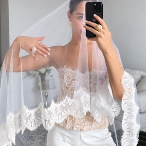 May include: A white bridal veil featuring a wide, intricate floral lace trim along the edges. The veil is made of sheer, lightweight tulle fabric. It is paired with a strapless, beige-toned corset top with lace detailing and white trousers.
