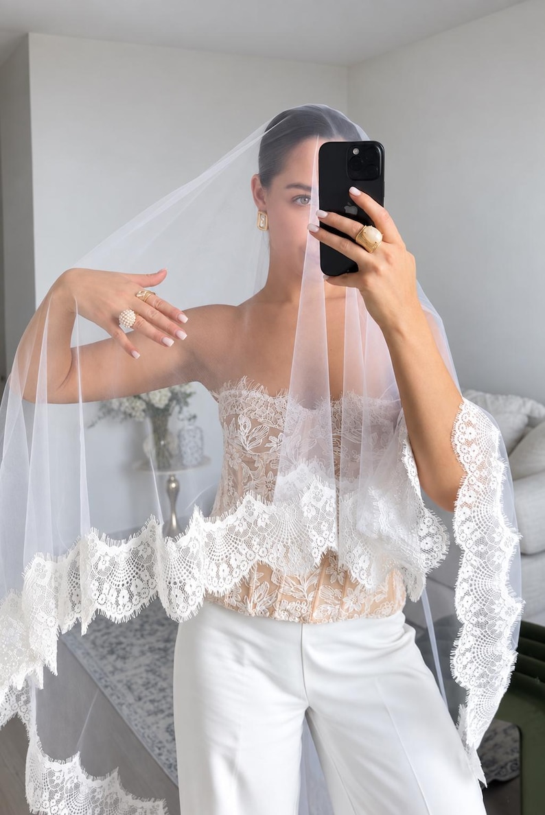 May include: A white bridal veil featuring a wide, intricate floral lace trim along the edges. The veil is made of sheer, lightweight tulle fabric. It is paired with a strapless, beige-toned corset top with lace detailing and white trousers.