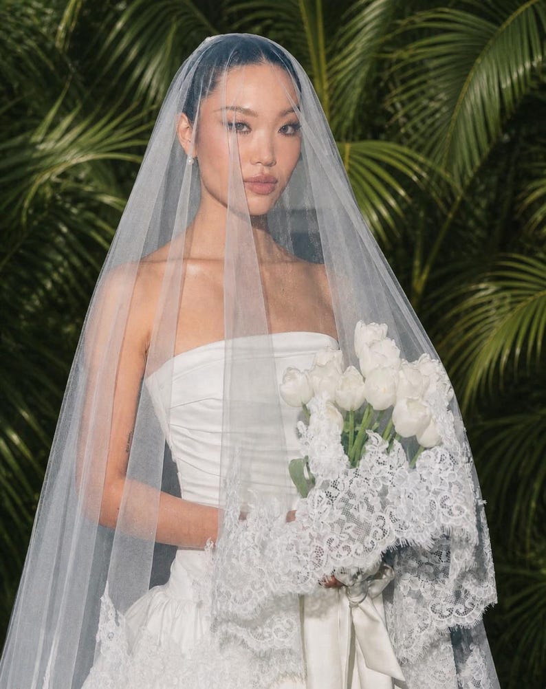 May include: A sheer white bridal veil featuring intricate lace trim along the edges, draped over a strapless white gown. The veil is long and translucent, partially covering a bouquet of white tulips held in front of the white dress.