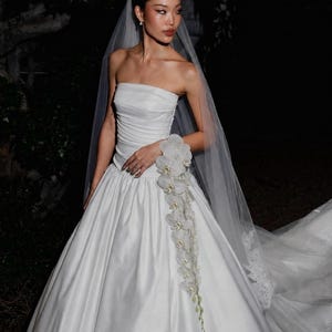 May include: A white strapless wedding gown with a ruched bodice and a full, floor-length skirt. A long, sheer veil is attached to the hair. A cascading arrangement of white orchid flowers is pinned to the side of the waistline of the dress.
