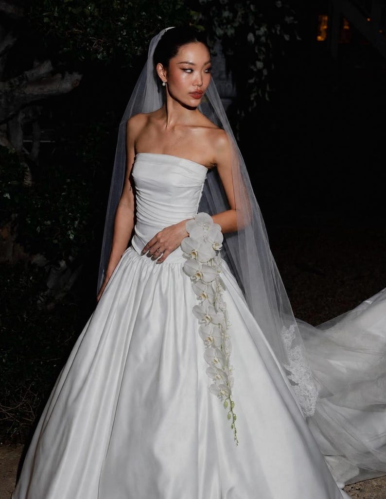 May include: A white strapless wedding gown with a ruched bodice and a full, floor-length skirt. A long, sheer veil is attached to the hair. A cascading arrangement of white orchid flowers is pinned to the side of the waistline of the dress.