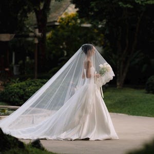 May include: A floor-length white strapless wedding gown with a full skirt and a long, sheer, floor-sweeping veil. The veil features delicate lace trim along the edges, and a bouquet of white flowers is held in the hand of the wearer.