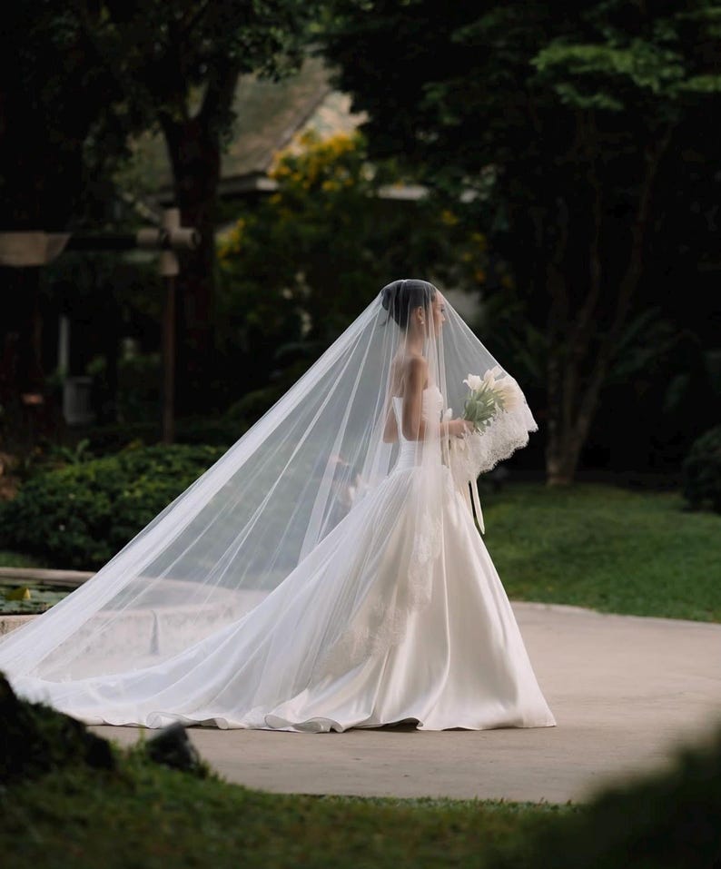 May include: A floor-length white strapless wedding gown with a full skirt and a long, sheer, floor-sweeping veil. The veil features delicate lace trim along the edges, and a bouquet of white flowers is held in the hand of the wearer.