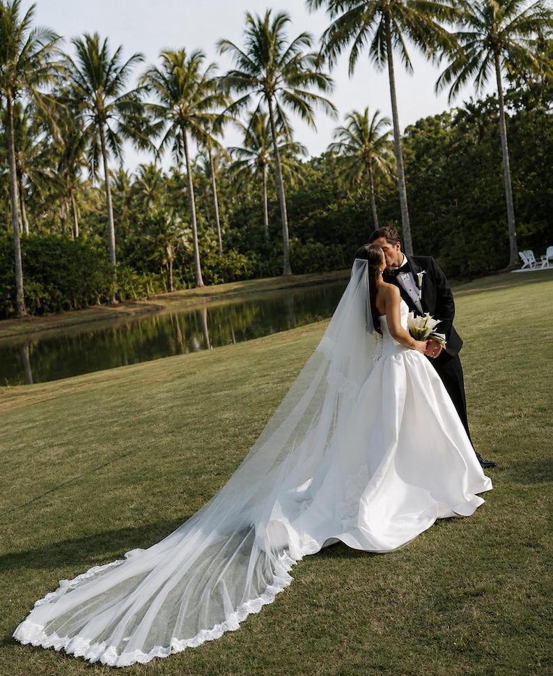 May include: A white strapless wedding gown with a full skirt and a long, sheer lace-trimmed veil, paired with a black tuxedo featuring a white shirt and bow tie. The attire is worn by two individuals standing together on a grassy lawn.