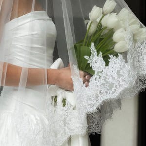 May include: A white strapless gown with a ruched bodice and a lace-trimmed hem, paired with a sheer tulle veil featuring intricate floral lace edges. A bouquet of white tulips is held, with the stems wrapped in a white ribbon with a bow.