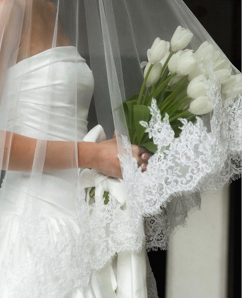 May include: A white strapless gown with a ruched bodice and a lace-trimmed hem, paired with a sheer tulle veil featuring intricate floral lace edges. A bouquet of white tulips is held, with the stems wrapped in a white ribbon with a bow.