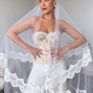 May include: A white bridal veil featuring a scalloped lace trim edge, paired with a strapless corset top adorned with intricate white floral embroidery over a nude base, and solid white wide-leg trousers, creating a layered ensemble.