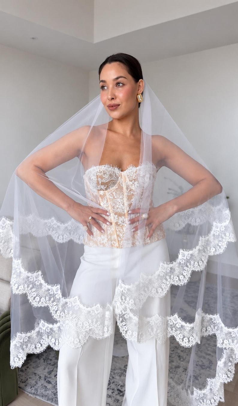 May include: A white bridal veil featuring a scalloped lace trim edge, paired with a strapless corset top adorned with intricate white floral embroidery over a nude base, and solid white wide-leg trousers, creating a layered ensemble.