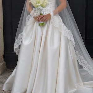 May include: A strapless ivory satin ball gown with a full skirt and a sweetheart neckline, paired with a long, sheer tulle veil featuring intricate white lace trim along the edges, while holding a bouquet of white calla lilies with green stems.