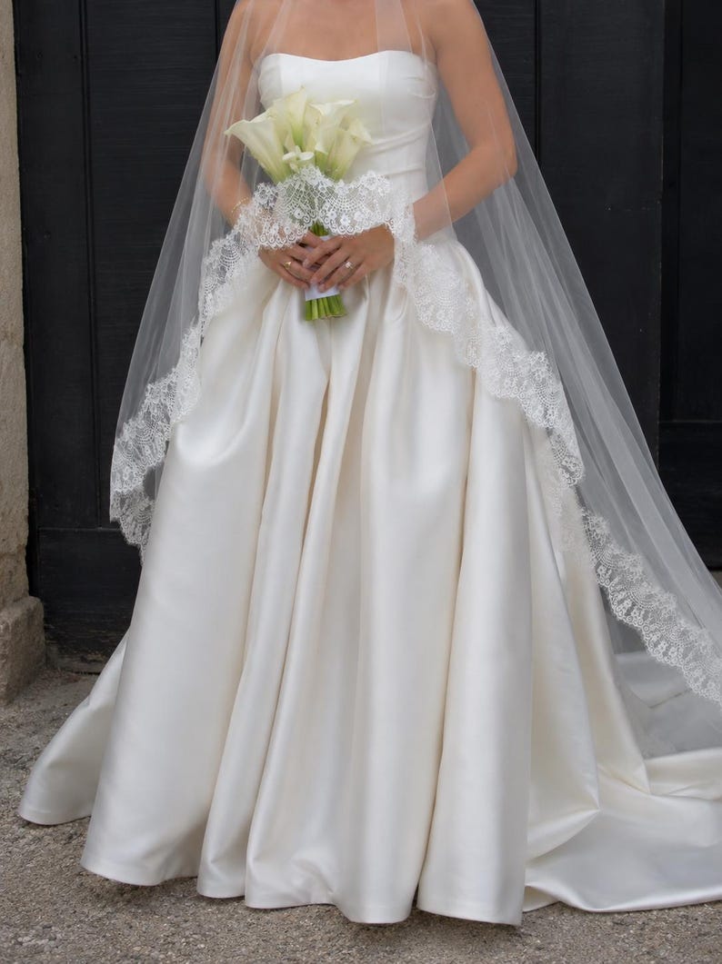 May include: A strapless ivory satin ball gown with a full skirt and a sweetheart neckline, paired with a long, sheer tulle veil featuring intricate white lace trim along the edges, while holding a bouquet of white calla lilies with green stems.