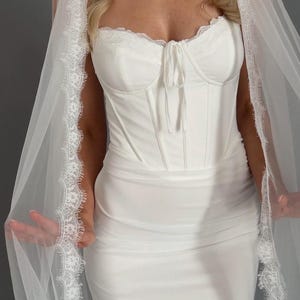 May include: A white bridal gown with a corset-style bodice, trimmed with lace, and a flowing skirt. A sheer veil with lace edging is draped over the dress. The dress is made of a smooth, opaque fabric, creating an elegant look for a wedding.