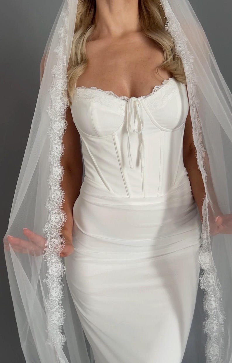 May include: A white bridal gown with a corset-style bodice, trimmed with lace, and a flowing skirt. A sheer veil with lace edging is draped over the dress. The dress is made of a smooth, opaque fabric, creating an elegant look for a wedding.