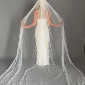 May include: A long, white bridal veil with a delicate lace trim. The veil is draped over a white wedding dress, creating a flowing, elegant look. The veil is made of a sheer, lightweight fabric.