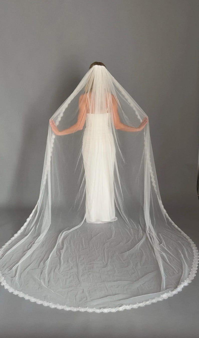 May include: A long, white bridal veil with a delicate lace trim. The veil is draped over a white wedding dress, creating a flowing, elegant look. The veil is made of a sheer, lightweight fabric.