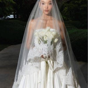 May include: A sheer white bridal veil featuring intricate lace trim along the edges, draped over a strapless white satin gown. The veil is paired with a bouquet of white tulips tied with a matching white satin ribbon, creating a cohesive look.