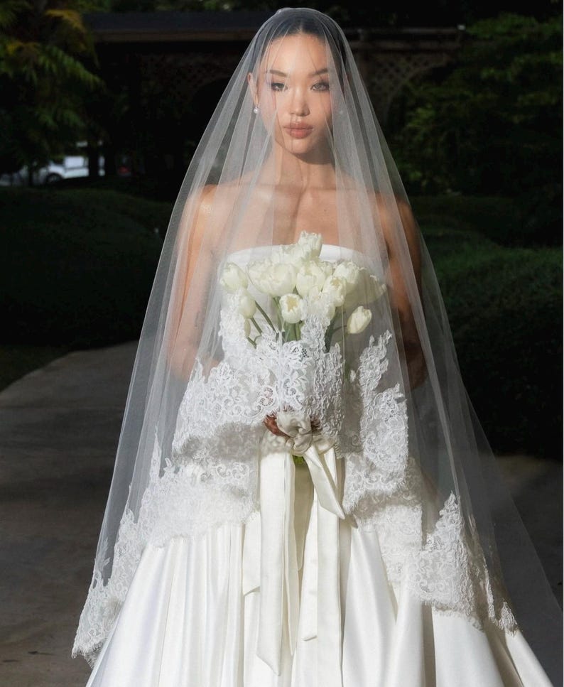 May include: A sheer white bridal veil featuring intricate lace trim along the edges, draped over a strapless white satin gown. The veil is paired with a bouquet of white tulips tied with a matching white satin ribbon, creating a cohesive look.