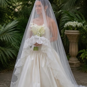 May include: A floor-length ivory wedding dress with a strapless bodice and a full, satin skirt. A sheer, white tulle veil with intricate white lace trim along the edges covers the head and shoulders. A bouquet of white calla lilies is held.