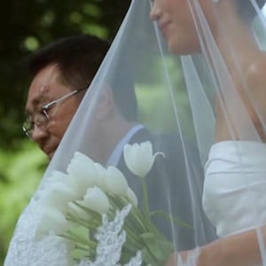 May include: A white strapless wedding dress paired with a long, sheer veil featuring intricate white lace trim along the edges. The ensemble is complemented by a bouquet of fresh white tulips held by the wearer, who also wears simple pearl earrings.