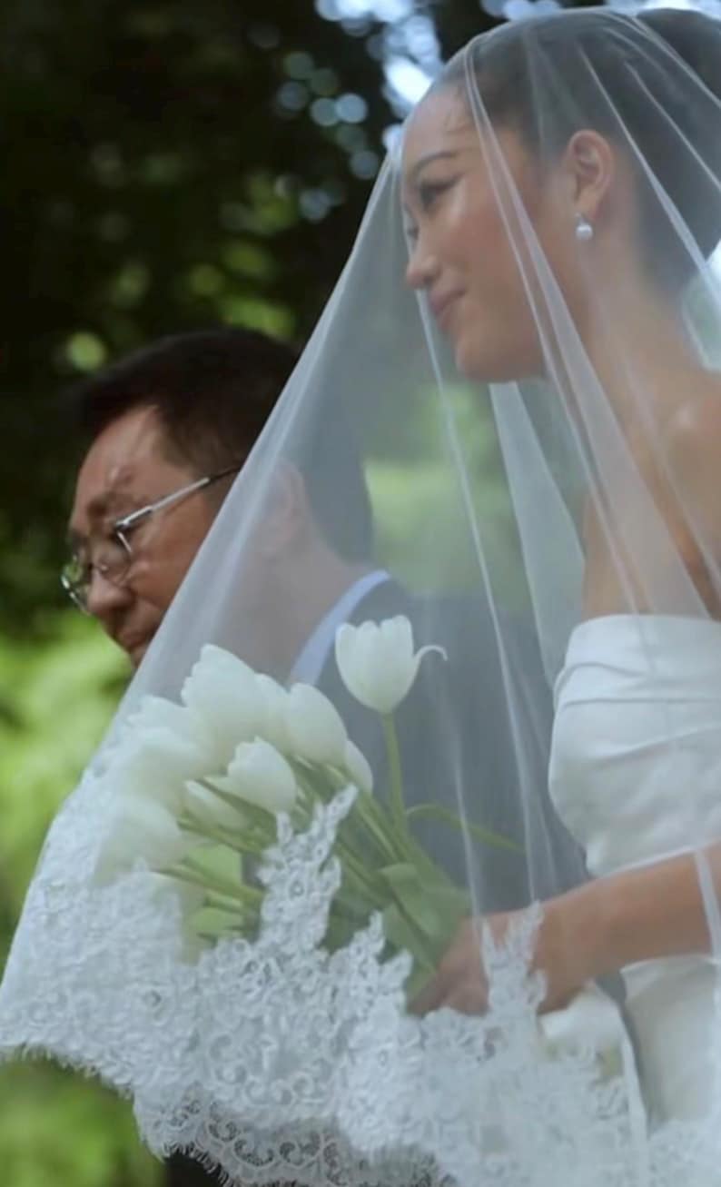 May include: A white strapless wedding dress paired with a long, sheer veil featuring intricate white lace trim along the edges. The ensemble is complemented by a bouquet of fresh white tulips held by the wearer, who also wears simple pearl earrings.