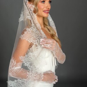May include: A white wedding veil with intricate lace detailing. The veil is draped over a white dress and matching lace gloves. The bride is smiling, with blonde hair and blue eyes. The background is a solid gray.