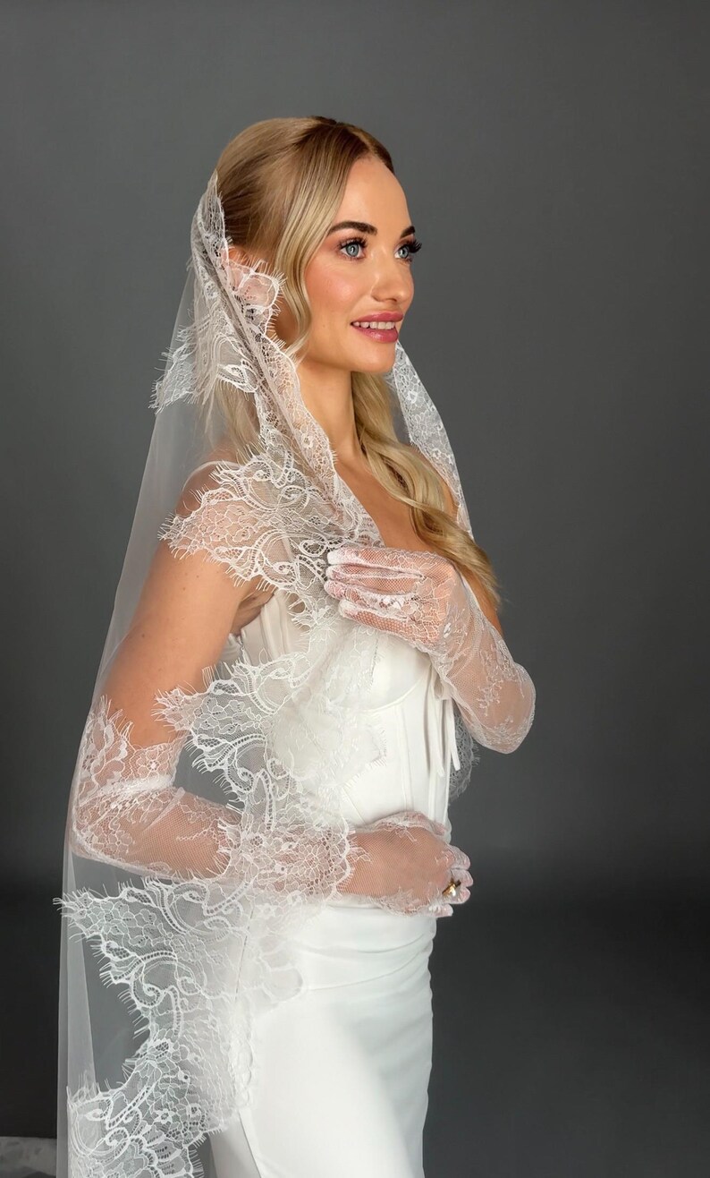 May include: A white wedding veil with intricate lace detailing. The veil is draped over a white dress and matching lace gloves. The bride is smiling, with blonde hair and blue eyes. The background is a solid gray.