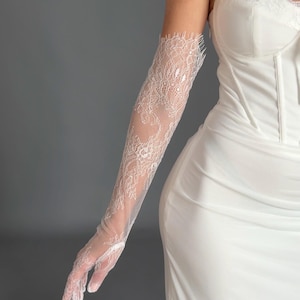 May include: White lace gloves with a delicate floral pattern. The gloves are long, extending up the arm, and are paired with a white dress. The lace is intricate, and the overall look is elegant and formal.