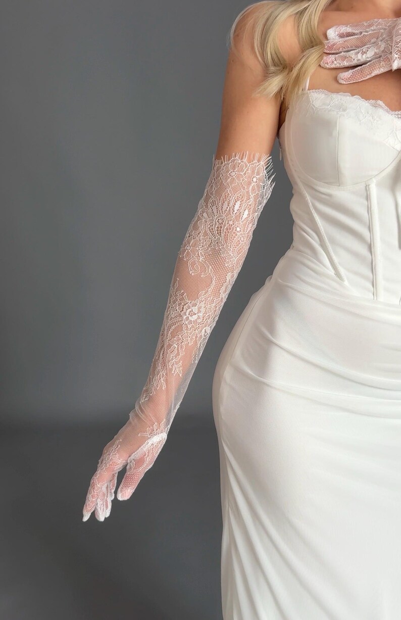 May include: White lace gloves with a delicate floral pattern. The gloves are long, extending up the arm, and are paired with a white dress. The lace is intricate, and the overall look is elegant and formal.