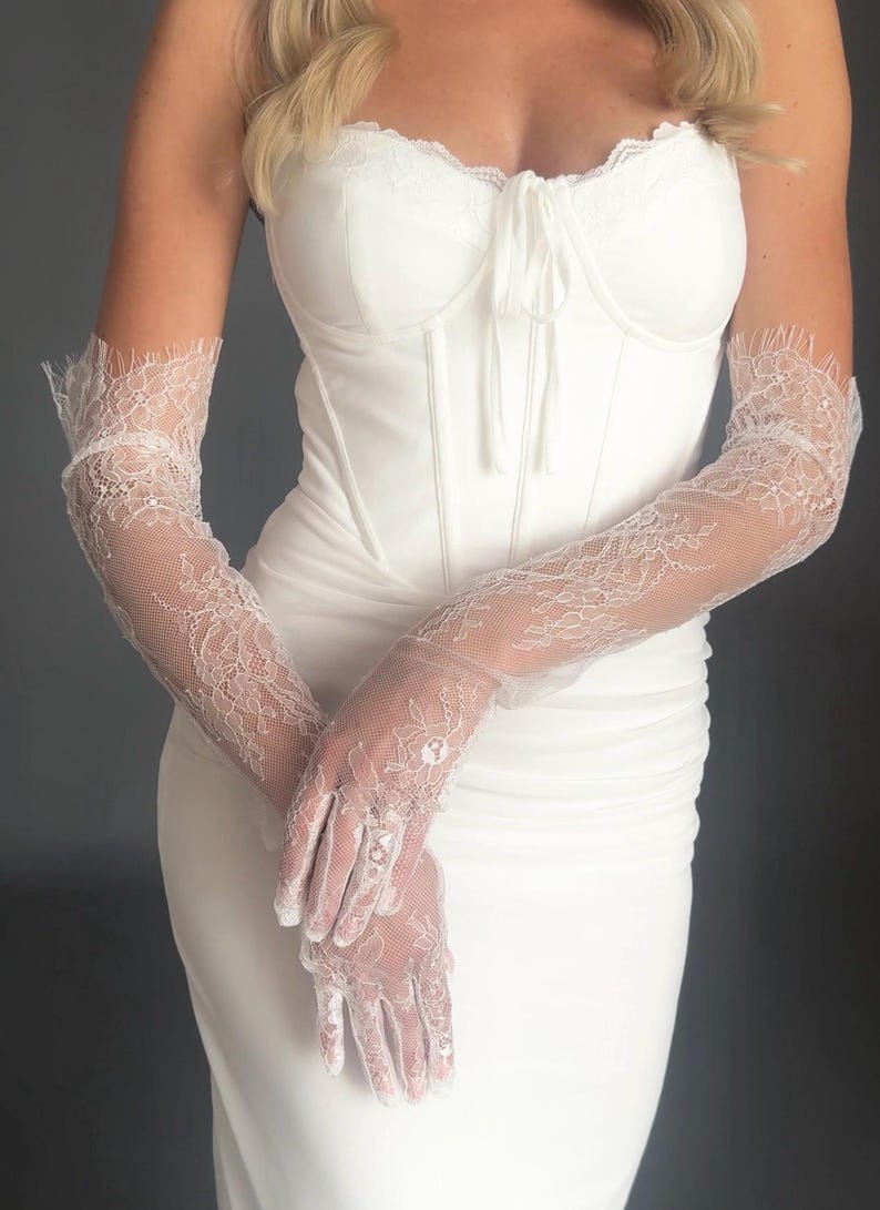 May include: A white, strapless corset-style bodice dress with a sweetheart neckline. The dress is paired with long, sheer white lace gloves. The gloves feature intricate floral lace patterns and extend to the upper arm, creating an elegant look.
