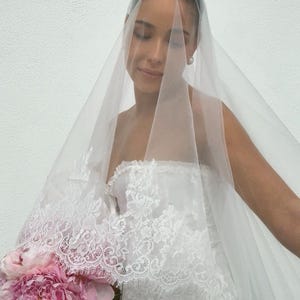 May include: A sheer white bridal veil featuring intricate floral lace trim along the edges, paired with a white strapless lace corset top and a bouquet of large pink peony flowers. The veil drapes over the head and shoulders in a soft, translucent layer.