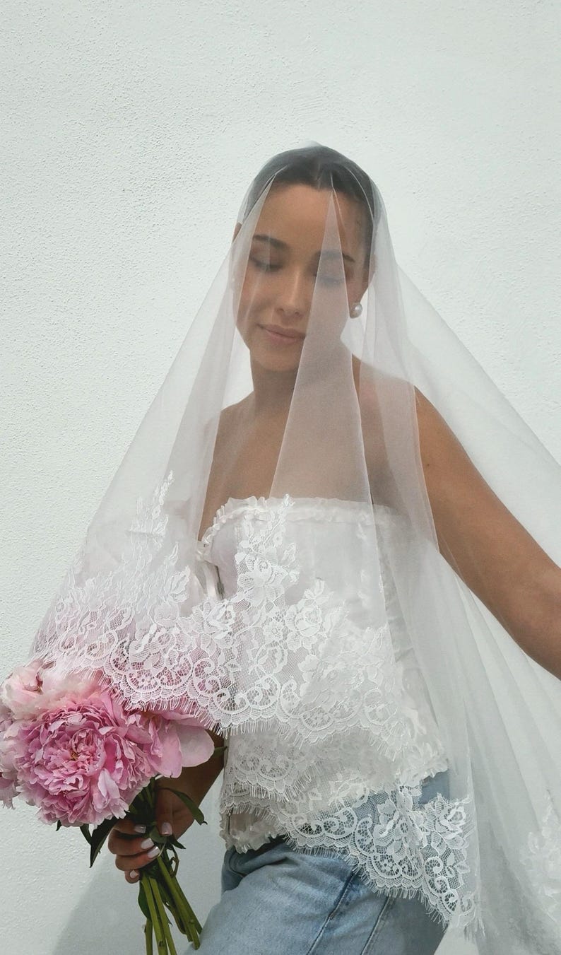 May include: A sheer white bridal veil featuring intricate floral lace trim along the edges, paired with a white strapless lace corset top and a bouquet of large pink peony flowers. The veil drapes over the head and shoulders in a soft, translucent layer.