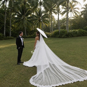 May include: A person in a white strapless wedding gown with a long, floor-sweeping lace-trimmed veil and a white parasol, standing near another person in a black tuxedo with a white shirt and black bow tie on a grassy lawn.