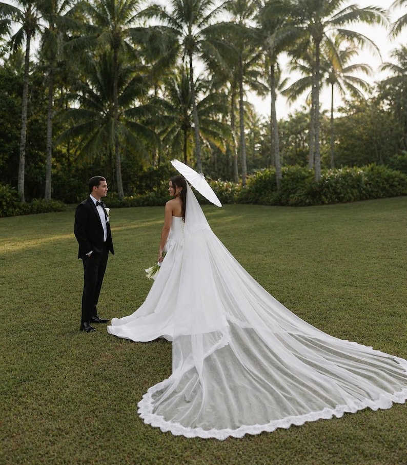 May include: A person in a white strapless wedding gown with a long, floor-sweeping lace-trimmed veil and a white parasol, standing near another person in a black tuxedo with a white shirt and black bow tie on a grassy lawn.