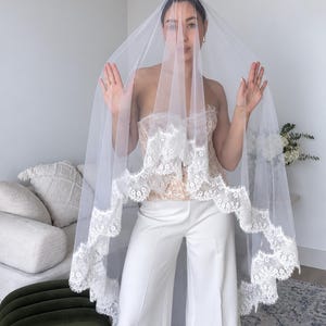 May include: A sheer white bridal veil featuring a scalloped lace trim with intricate floral embroidery along the edges. The veil is made of lightweight tulle fabric that drapes over the head and shoulders, showcasing a delicate and detailed lace border.