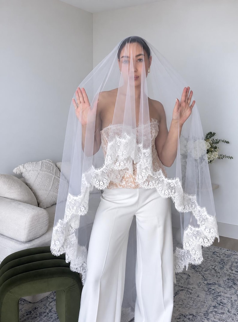May include: A sheer white bridal veil featuring a scalloped lace trim with intricate floral embroidery along the edges. The veil is made of lightweight tulle fabric that drapes over the head and shoulders, showcasing a delicate and detailed lace border.