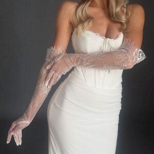 May include: A white wedding dress with a fitted bodice and a flowing skirt. The dress is paired with sheer, long lace gloves. The gloves feature intricate floral lace patterns. The overall look is elegant and bridal.
