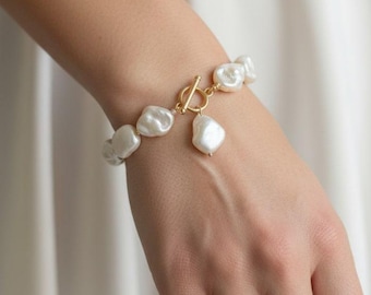 May include: A gold-toned bracelet featuring irregular, white baroque pearls. The bracelet has a toggle clasp and a dangling pearl charm. The jewelry is displayed on a wrist, with a simple gold ring on a finger.