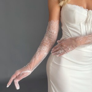 May include: Elegant white lace gloves, extending to the upper arm, paired with a white corset-style top and a flowing white dress. The gloves feature intricate floral lace patterns, adding a touch of sophistication to the bridal or formal attire.