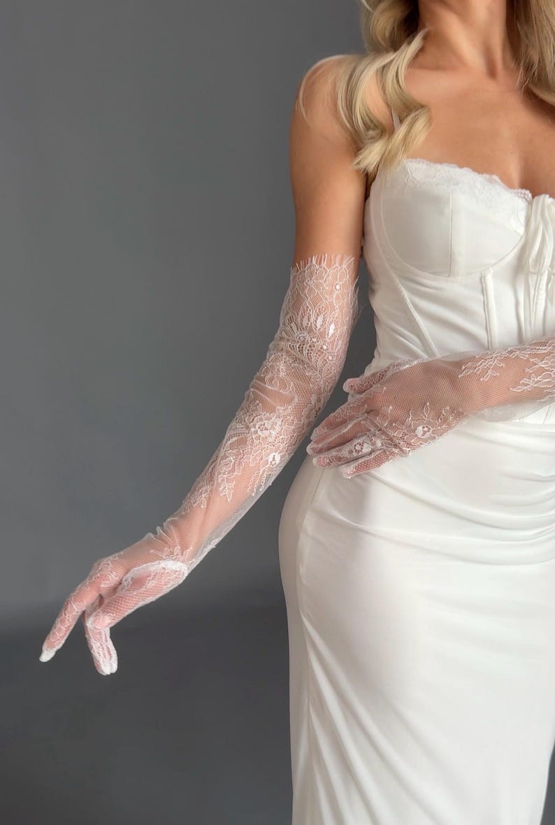 May include: Elegant white lace gloves, extending to the upper arm, paired with a white corset-style top and a flowing white dress. The gloves feature intricate floral lace patterns, adding a touch of sophistication to the bridal or formal attire.