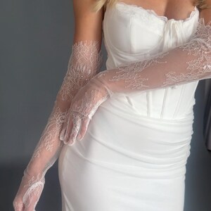 May include: White bridal gown with a sweetheart neckline and a fitted bodice. The dress is paired with long, sheer lace gloves. The gloves feature intricate floral patterns. The overall look is elegant and suitable for a wedding or formal event.