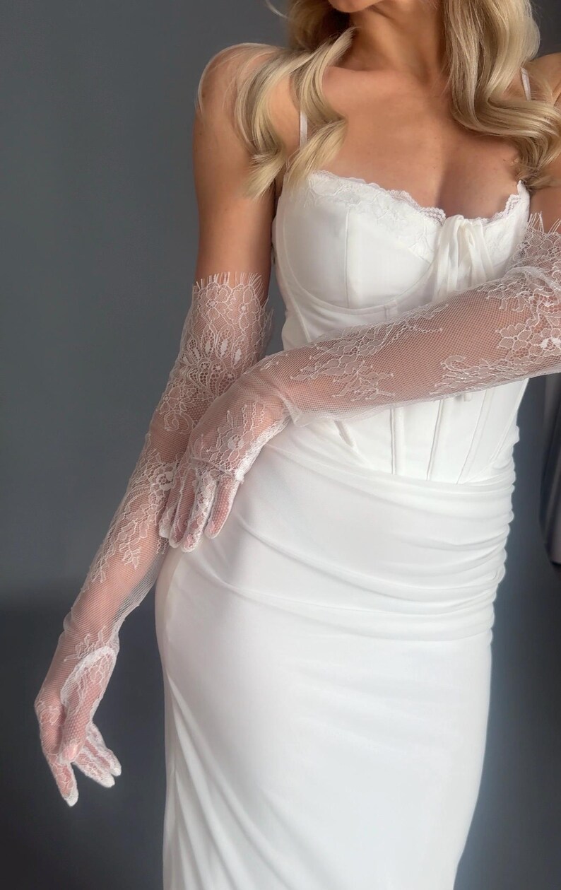 May include: White bridal gown with a sweetheart neckline and a fitted bodice. The dress is paired with long, sheer lace gloves. The gloves feature intricate floral patterns. The overall look is elegant and suitable for a wedding or formal event.