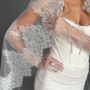 May include: A white wedding dress with a fitted bodice and a flowing skirt. The dress is paired with a long, lace-trimmed veil and matching lace gloves. A gold ring with a pearl is visible.
