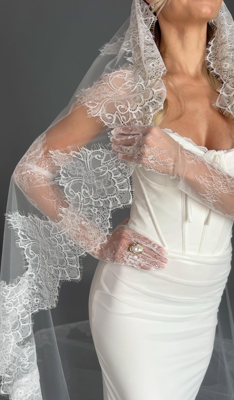 May include: A white wedding dress with a fitted bodice and a flowing skirt. The dress is paired with a long, lace-trimmed veil and matching lace gloves. A gold ring with a pearl is visible.