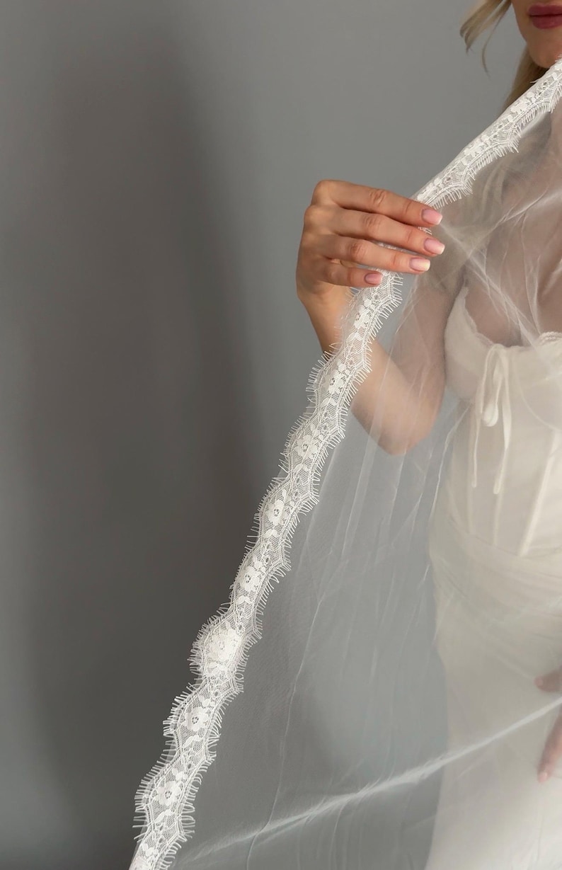 May include: A sheer white wedding veil with a detailed lace trim. The veil is held up, displaying the intricate floral pattern of the lace. The veil is worn with a white dress, creating a classic bridal look.